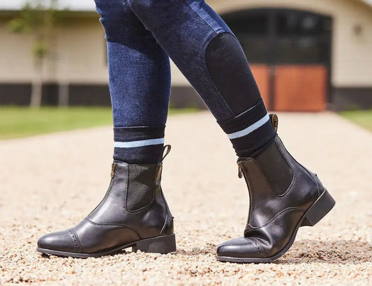Person wearing black paddock boots and breeches with a stable in the background