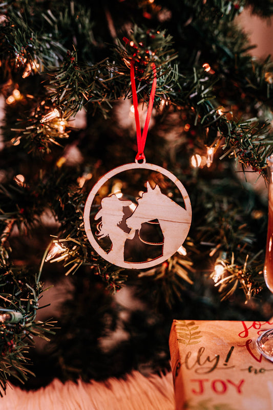 A round, unfinished birch wood Christmas ornament featuring an engraved depiction of a girl and a horse, with a red ribbon for hanging, displayed on a Christmas tree.